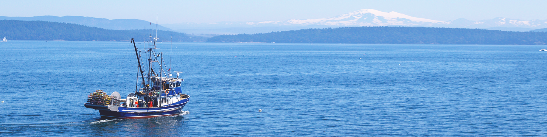 A small commercial fishing boat moves through calm blue water, with tree-lined islands and distant snow-capped mountains under a clear sky.