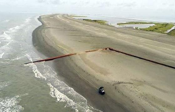 An aerial view of a sandy beach with waves gently crashing onto the shore. A long, narrow strip of land extends into the distance, bordered by water on both sides. A rust-colored pipeline runs diagonally across the beach from the ocean to an inland area. A small vehicle is parked near the pipeline on the sand. The sky is overcast with light clouds.