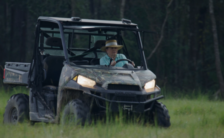 A person wearing a wide‑brimmed hat drives a camouflaged utility vehicle through tall grass, with forest trees in the background.