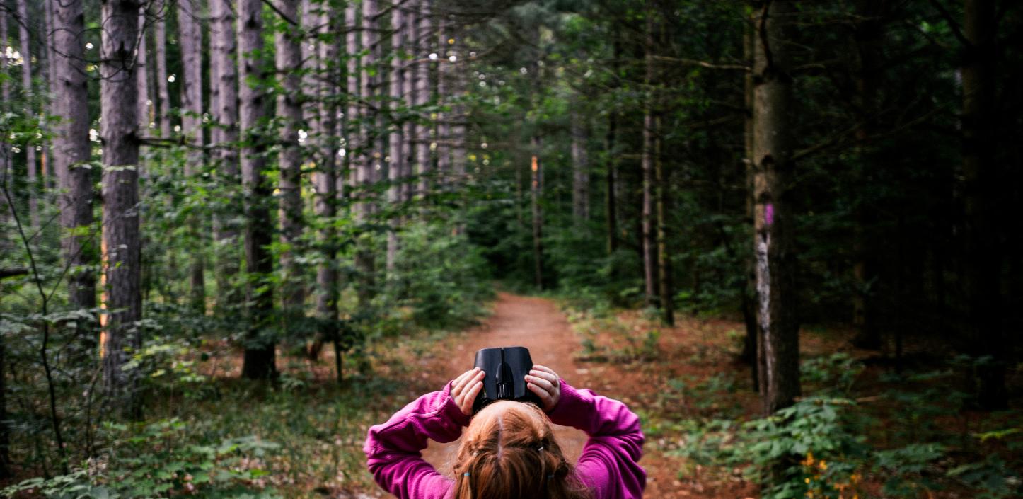 a young girl standing on a forest path looks up at the tree canopy through binoculars