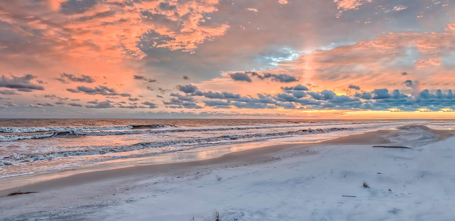 A wide, tranquil beach at sunrise with soft waves rolling in beneath a sky glowing with pink and orange clouds.