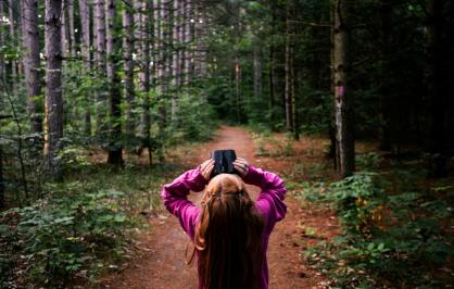 a young girl standing on a forest path looks up at the tree canopy through binoculars