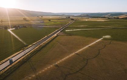 Aerial view of a rural landscape at sunset, showing wide open farm fields divided by a straight two‑lane road. A blue truck travels along the road, and several irrigation systems spray water across the fields. Rolling hills and a bright, low sun appear in the distance.