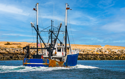 Commercial fishing boat heading out through a coastal inlet, with a rocky jetty and sandy dunes under a blue sky in the background.