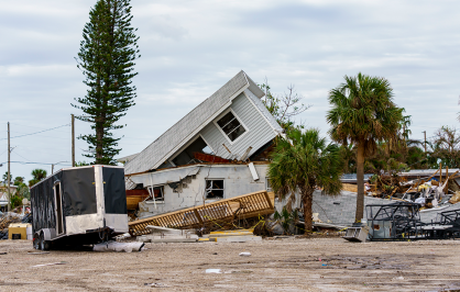 A storm-damaged coastal neighborhood with a collapsed home at the center of the scene. The roof of the house has partially caved in and rests at an angle atop shattered walls. Debris, broken structures, and uprooted vegetation are scattered across the property. A black enclosed trailer sits in the foreground, while palm trees and other damaged buildings stand in the background beneath a cloudy, overcast sky.