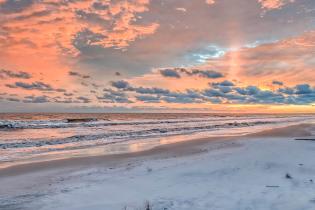 A wide, tranquil beach at sunrise with soft waves rolling in beneath a sky glowing with pink and orange clouds.