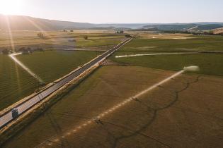 Aerial view of a rural landscape at sunset, showing wide open farm fields divided by a straight two‑lane road. A blue truck travels along the road, and several irrigation systems spray water across the fields. Rolling hills and a bright, low sun appear in the distance.