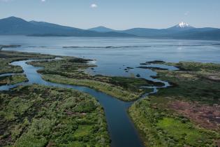 Aerial view of a winding river flowing through a lush green wetland toward a broad lake, with forested mountains and a snow‑capped peak in the distance under a clear blue sky.