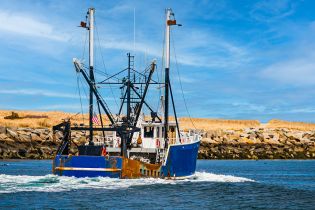 Commercial fishing boat heading out through a coastal inlet, with a rocky jetty and sandy dunes under a blue sky in the background.