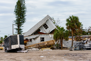 A storm-damaged coastal neighborhood with a collapsed home at the center of the scene. The roof of the house has partially caved in and rests at an angle atop shattered walls. Debris, broken structures, and uprooted vegetation are scattered across the property. A black enclosed trailer sits in the foreground, while palm trees and other damaged buildings stand in the background beneath a cloudy, overcast sky.
